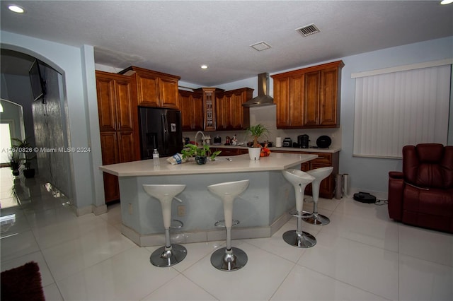 kitchen with wall chimney exhaust hood, a textured ceiling, black refrigerator with ice dispenser, a kitchen bar, and a center island with sink