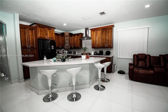 kitchen featuring wall chimney exhaust hood, a kitchen island with sink, black refrigerator with ice dispenser, and a breakfast bar area