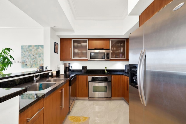 kitchen with appliances with stainless steel finishes, a tray ceiling, dark stone counters, crown molding, and sink