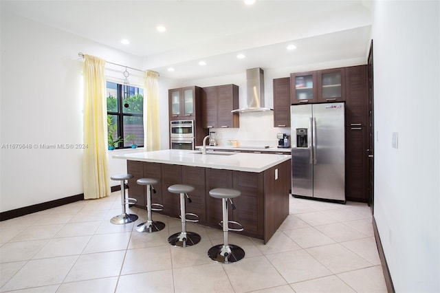 kitchen with sink, a kitchen island with sink, wall chimney range hood, stainless steel appliances, and a kitchen breakfast bar