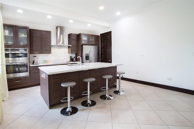 kitchen featuring appliances with stainless steel finishes, a breakfast bar, light tile patterned floors, a center island with sink, and wall chimney range hood