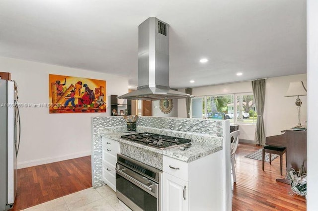 kitchen featuring light wood-type flooring, light stone counters, white cabinetry, island exhaust hood, and appliances with stainless steel finishes