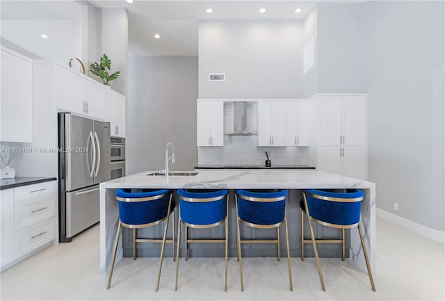 kitchen featuring sink, a center island with sink, wall chimney range hood, white cabinetry, and stainless steel refrigerator