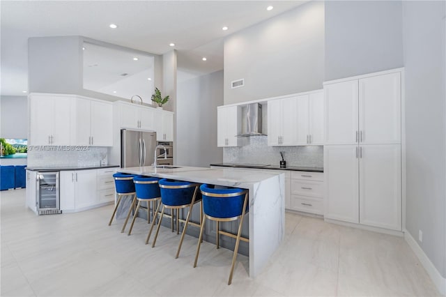 kitchen with wine cooler, stainless steel fridge, a center island with sink, wall chimney range hood, and white cabinetry