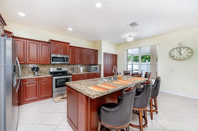 kitchen with stainless steel appliances, a sink, decorative backsplash, light tile patterned floors, and a center island with sink