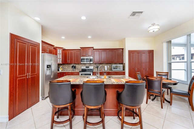 kitchen featuring appliances with stainless steel finishes, sink, backsplash, light stone counters, and a center island with sink