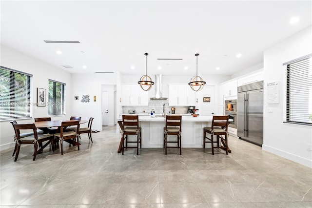 kitchen featuring wall chimney range hood, appliances with stainless steel finishes, white cabinetry, decorative light fixtures, and a center island with sink