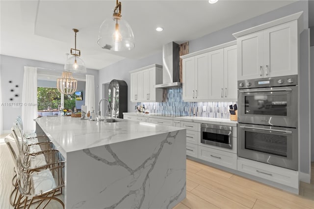 kitchen with white cabinetry, hanging light fixtures, double oven, wall chimney range hood, and a large island