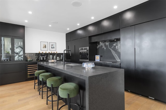 kitchen with wine cooler, a kitchen island with sink, tasteful backsplash, light hardwood / wood-style flooring, and oven