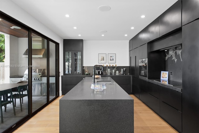 kitchen featuring sink, a kitchen island with sink, light hardwood / wood-style flooring, oven, and wall chimney range hood