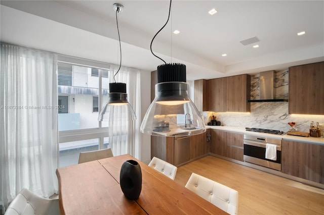 kitchen with wall chimney range hood, light hardwood / wood-style flooring, sink, stainless steel appliances, and hanging light fixtures