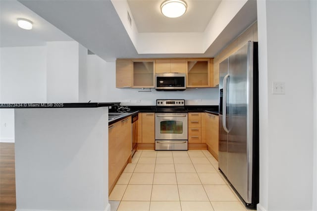 kitchen with light brown cabinetry, appliances with stainless steel finishes, light tile patterned flooring, kitchen peninsula, and a tray ceiling