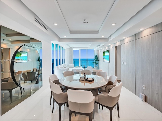 dining room featuring a water view, a tray ceiling, a wall of windows, wooden walls, and light tile patterned floors