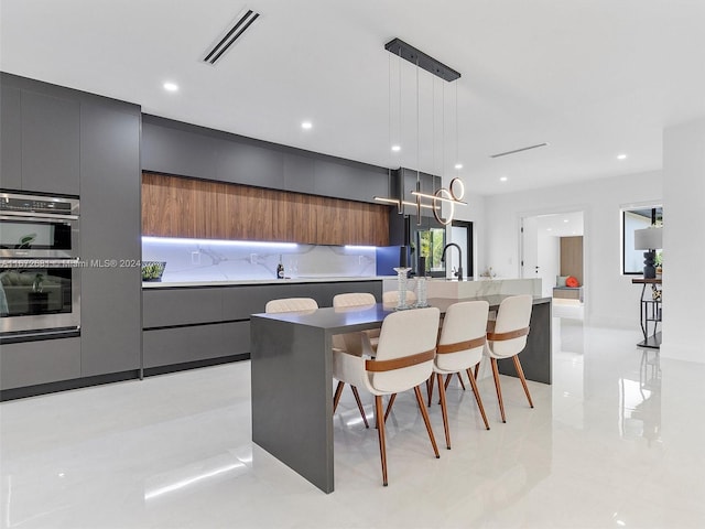 kitchen featuring double oven, an island with sink, decorative light fixtures, and a breakfast bar