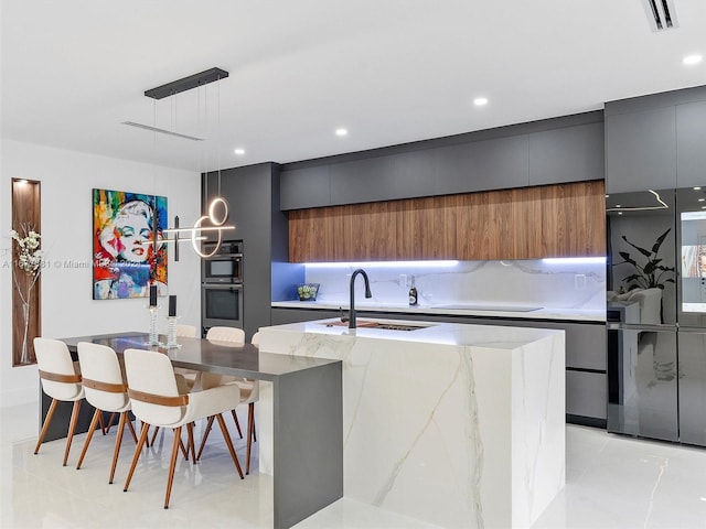 kitchen featuring pendant lighting, stainless steel double oven, a center island with sink, sink, and light stone countertops