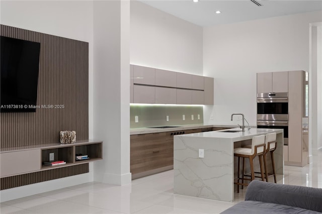 kitchen with sink, a breakfast bar, a high ceiling, a center island with sink, and stainless steel double oven