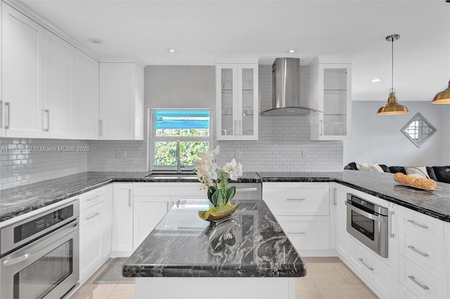 kitchen with hanging light fixtures, glass insert cabinets, a sink, wall chimney range hood, and oven