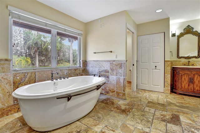 bathroom featuring a soaking tub, a wainscoted wall, stone tile flooring, vanity, and tile walls