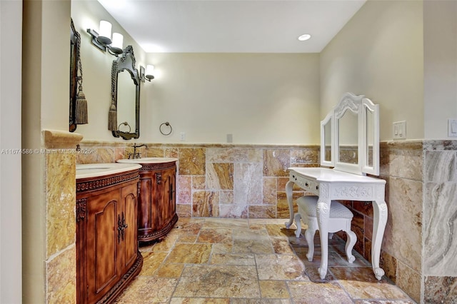 full bath with tile walls, a wainscoted wall, two vanities, stone tile flooring, and a sink