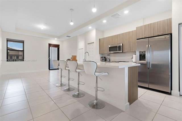 kitchen with a breakfast bar, a kitchen island with sink, light tile patterned floors, appliances with stainless steel finishes, and a tray ceiling