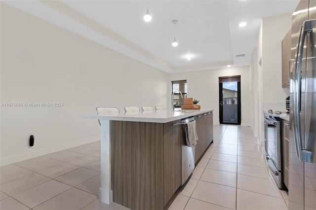 kitchen featuring decorative light fixtures, a tray ceiling, a kitchen island with sink, light tile patterned flooring, and appliances with stainless steel finishes