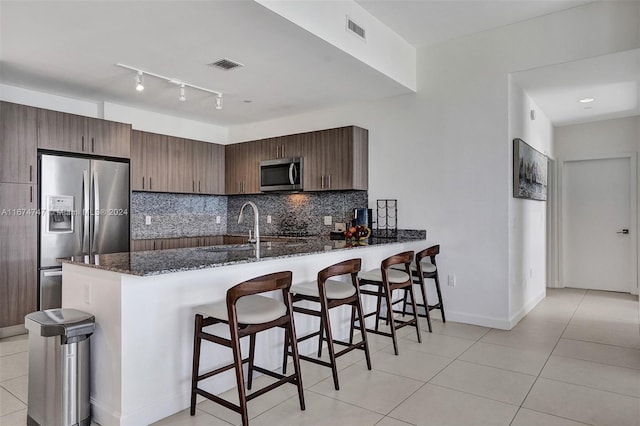 kitchen featuring dark brown cabinetry, stainless steel appliances, tasteful backsplash, dark stone countertops, and kitchen peninsula