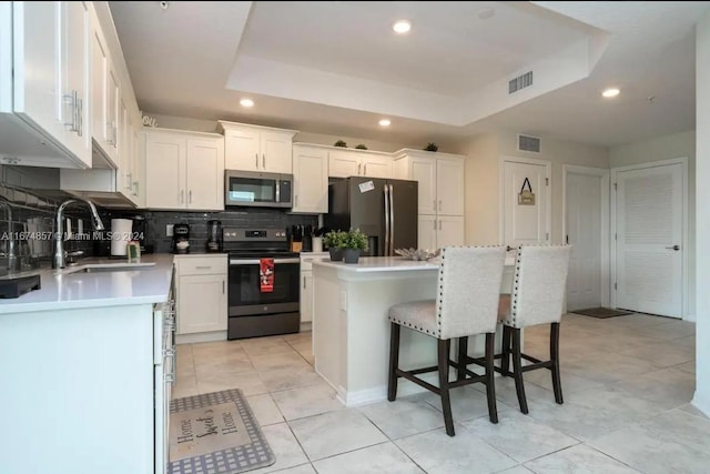 kitchen with a tray ceiling, a kitchen island, sink, stainless steel appliances, and white cabinetry