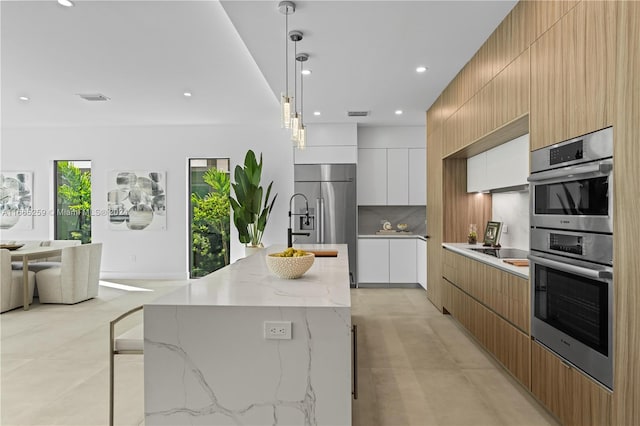 kitchen with pendant lighting, light stone counters, an island with sink, appliances with stainless steel finishes, and white cabinetry