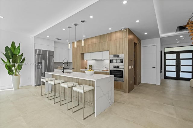 kitchen featuring decorative backsplash, a kitchen island with sink, stainless steel appliances, white cabinetry, and decorative light fixtures