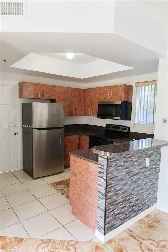 kitchen with appliances with stainless steel finishes, a tray ceiling, kitchen peninsula, dark stone countertops, and light tile patterned floors