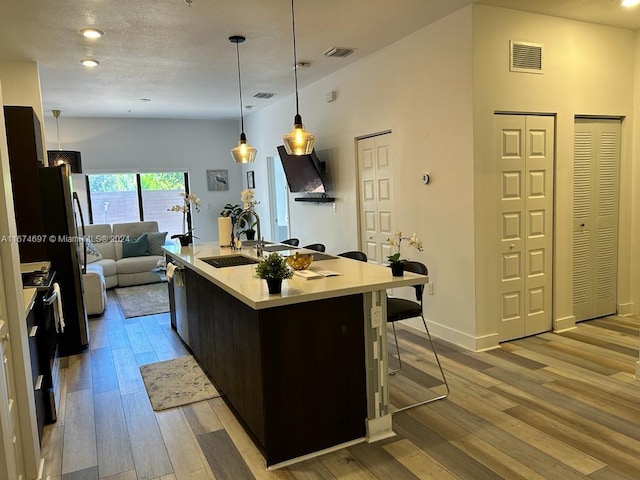 kitchen with light wood-type flooring, stainless steel stove, a center island with sink, sink, and hanging light fixtures