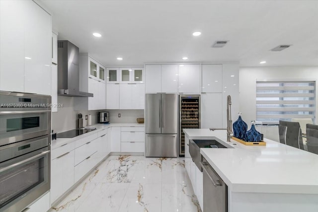 kitchen featuring a kitchen island with sink, stainless steel appliances, sink, wall chimney range hood, and white cabinetry