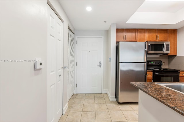 kitchen featuring light tile patterned floors, appliances with stainless steel finishes, brown cabinets, decorative backsplash, and dark stone countertops