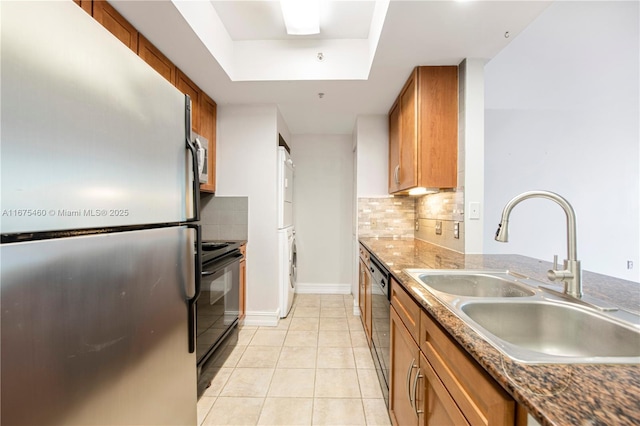 kitchen featuring a raised ceiling, brown cabinetry, stacked washer and clothes dryer, black appliances, and a sink