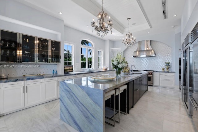 kitchen featuring wall chimney range hood, decorative backsplash, white cabinets, a center island with sink, and light stone counters