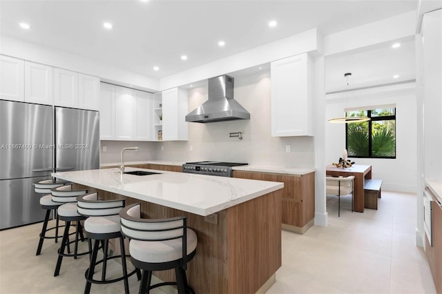 kitchen with wall chimney range hood, sink, stainless steel appliances, an island with sink, and white cabinetry