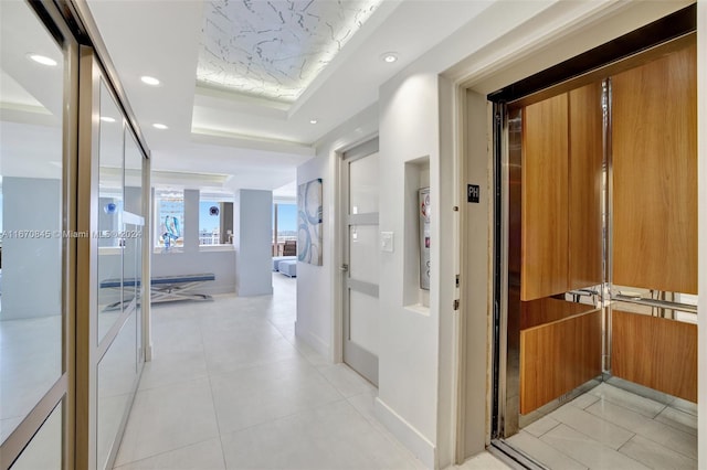 hallway featuring a tray ceiling, light tile patterned floors, and elevator