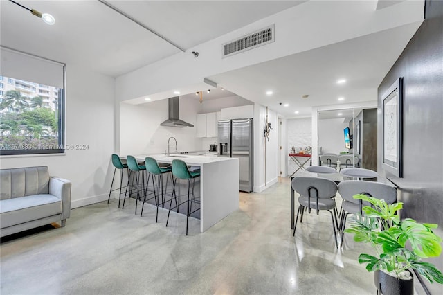 kitchen with wall chimney range hood, a breakfast bar, kitchen peninsula, white cabinets, and stainless steel fridge with ice dispenser