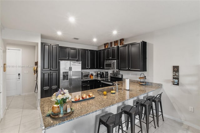 kitchen featuring appliances with stainless steel finishes, light stone countertops, dark cabinets, a peninsula, and a kitchen bar