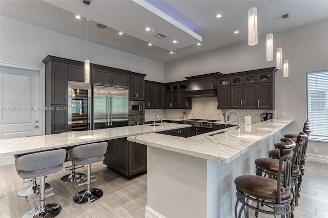 kitchen with kitchen peninsula, decorative light fixtures, built in appliances, custom range hood, and a breakfast bar