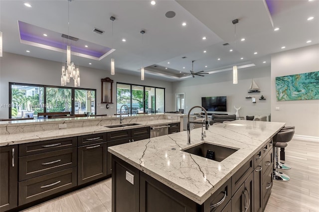 kitchen featuring a large island with sink, light stone counters, a raised ceiling, sink, and decorative light fixtures