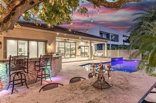 pool at dusk featuring exterior bar, ceiling fan, and a patio