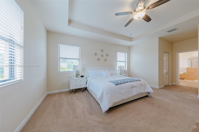 carpeted bedroom with a tray ceiling, ensuite bath, multiple windows, and ceiling fan