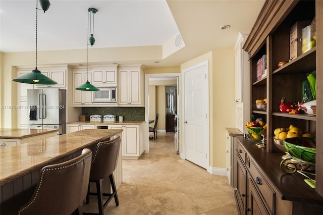 kitchen with decorative backsplash, hanging light fixtures, appliances with stainless steel finishes, light stone countertops, and cream cabinetry