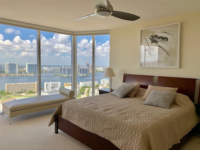 carpeted bedroom with a water view, ceiling fan, multiple windows, and a wall of windows