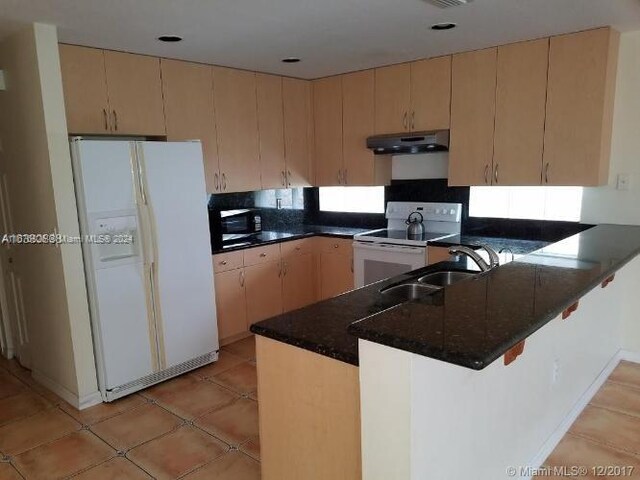 kitchen with light brown cabinets, kitchen peninsula, dark stone counters, sink, and white appliances