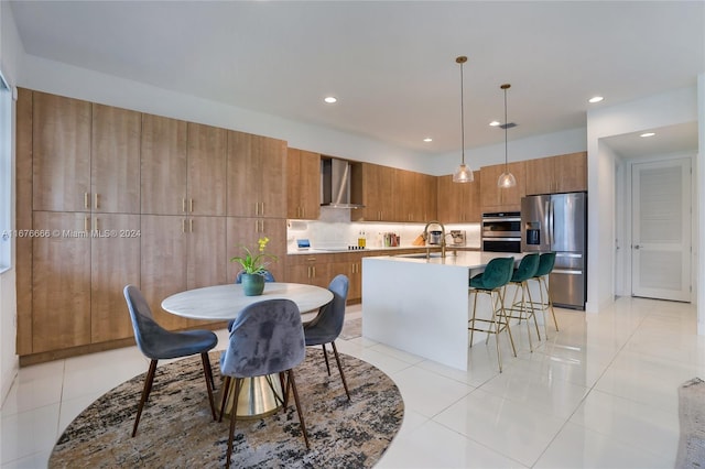 kitchen with wall chimney range hood, an island with sink, stainless steel appliances, decorative light fixtures, and light tile patterned floors