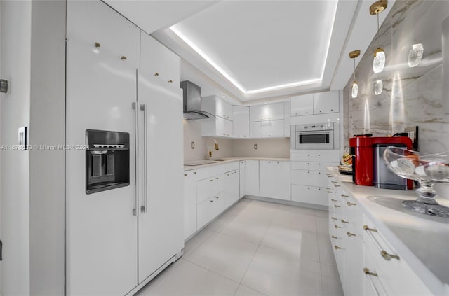 kitchen with white appliances, wall chimney range hood, a raised ceiling, white cabinetry, and pendant lighting