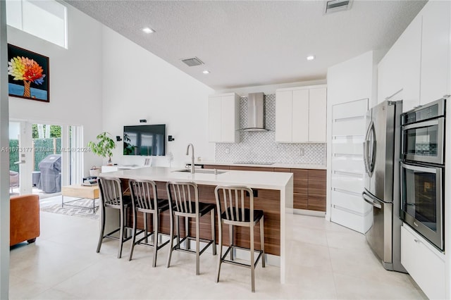 kitchen with appliances with stainless steel finishes, wall chimney exhaust hood, sink, white cabinetry, and an island with sink