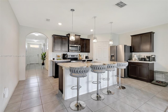 kitchen with light tile patterned floors, appliances with stainless steel finishes, an island with sink, dark brown cabinetry, and decorative light fixtures